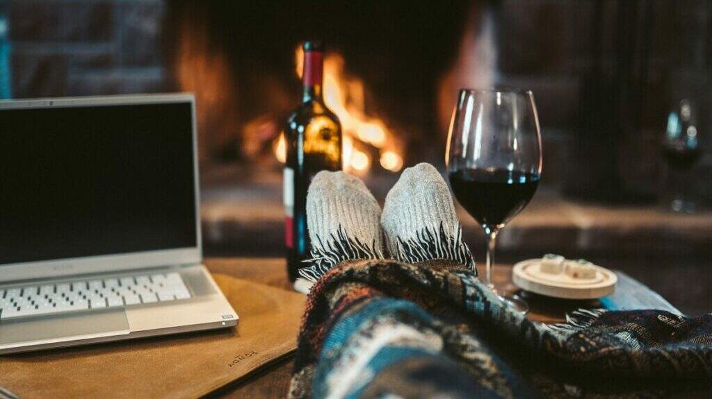 Pessoa com os pés em cima de uma mesa. Na mesa, uma taça de vinho e um notebook. Ao fundo, um cenário típico de inverno com lareira acesa